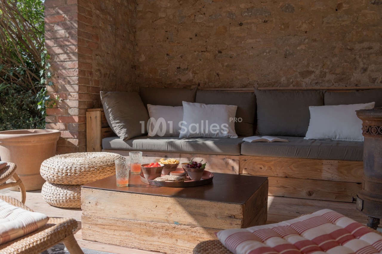 Terrasse en pierre avec canapé, coussins, table basse en bois et collations, éclairée par la lumière naturelle.