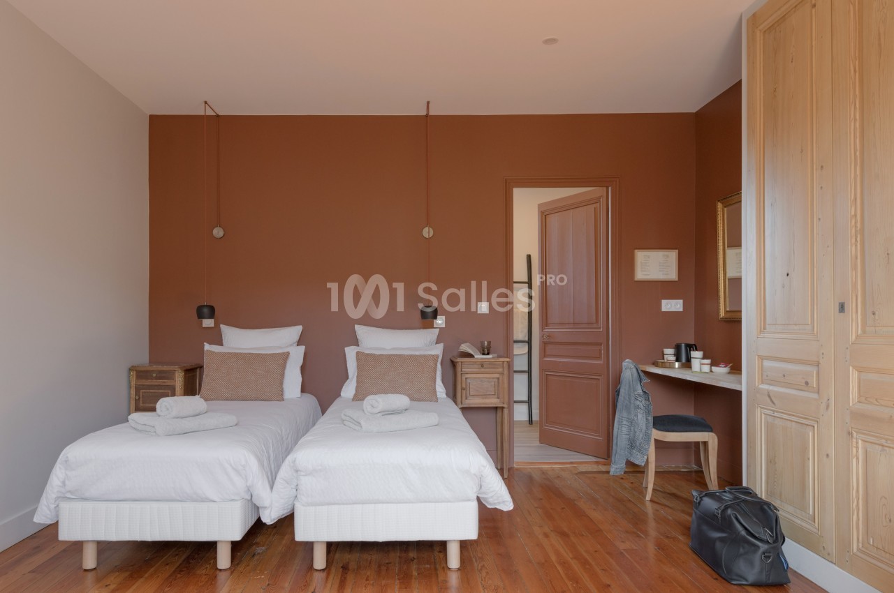 Chambre avec deux lits simples, murs marron, parquet en bois, bureau avec chaise et porte ouverte sur une autre pièce.
