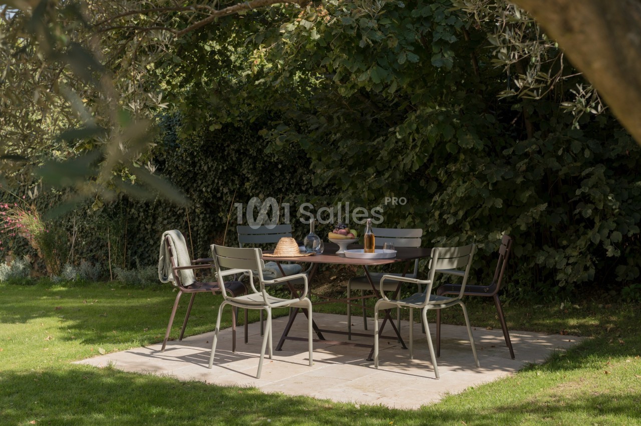 Table de jardin entourée de chaises sur une terrasse en pierre, sous des arbres, avec des plats et boissons disposés.