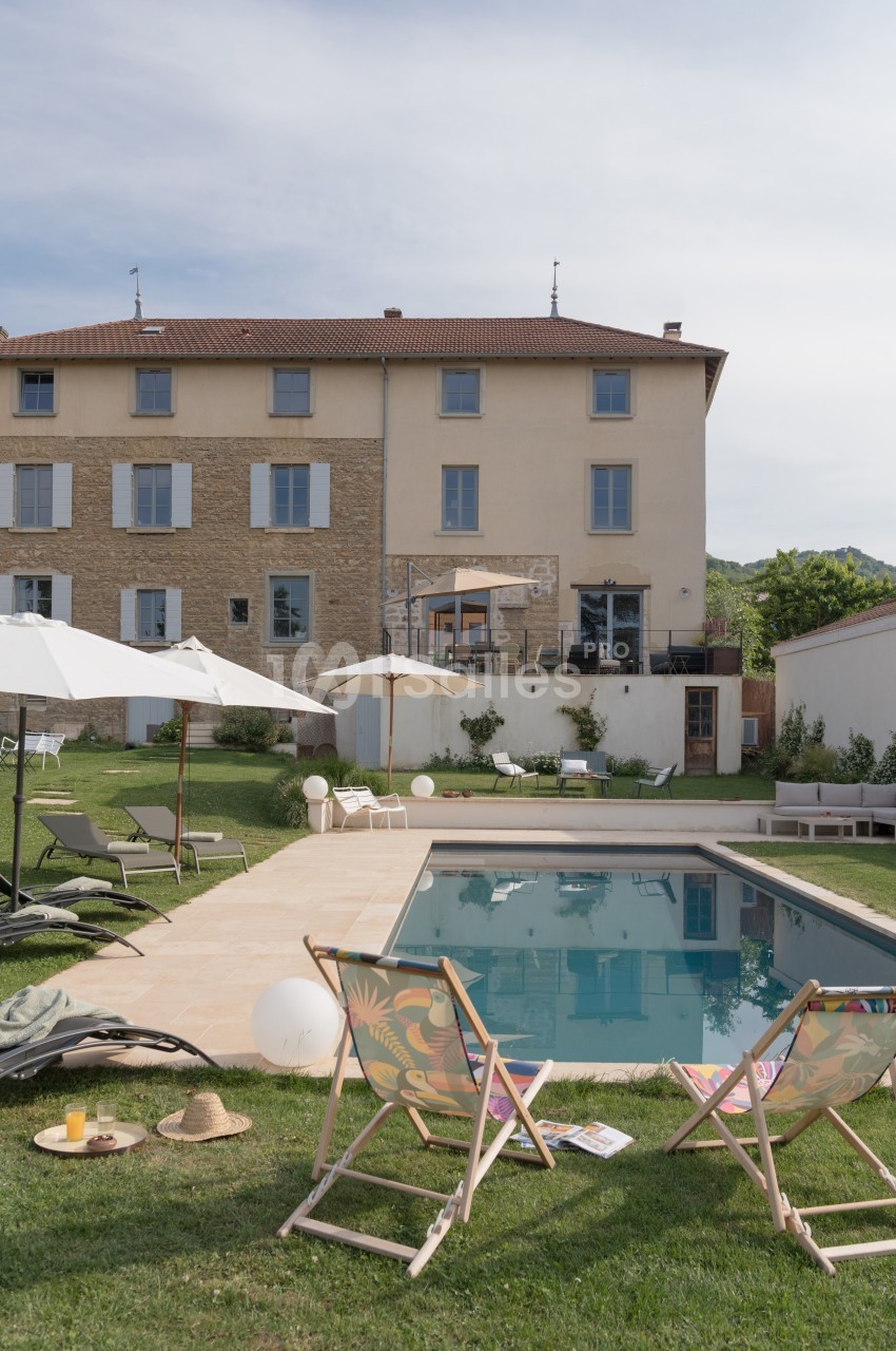 Jardin avec piscine, transats, parasols et maison en arrière-plan sous un ciel légèrement nuageux.