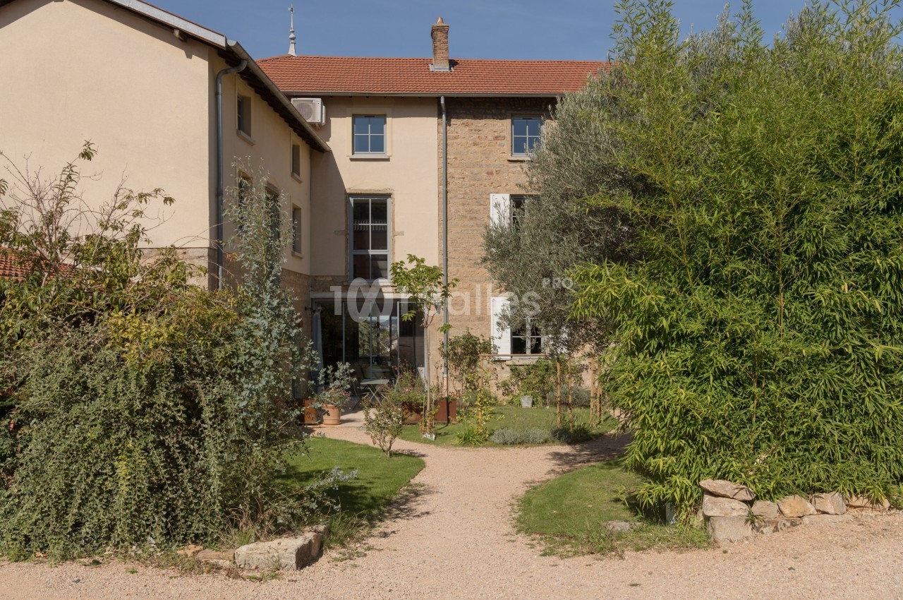 Façade d'une maison en pierre avec toit en tuiles, entourée de végétation et d'un chemin en gravier.