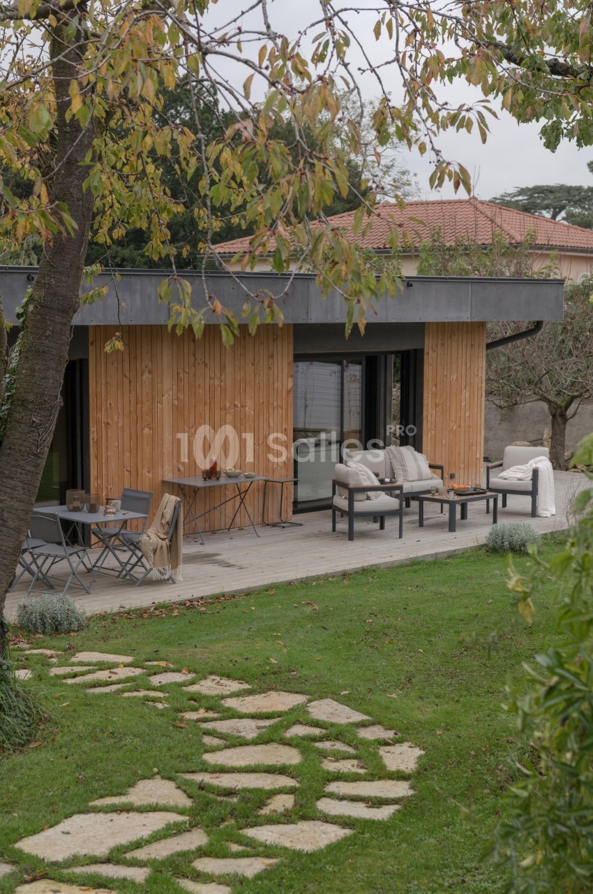 Terrasse aménagée avec mobilier de jardin devant une maison en bois, entourée de pelouse et d'arbres.