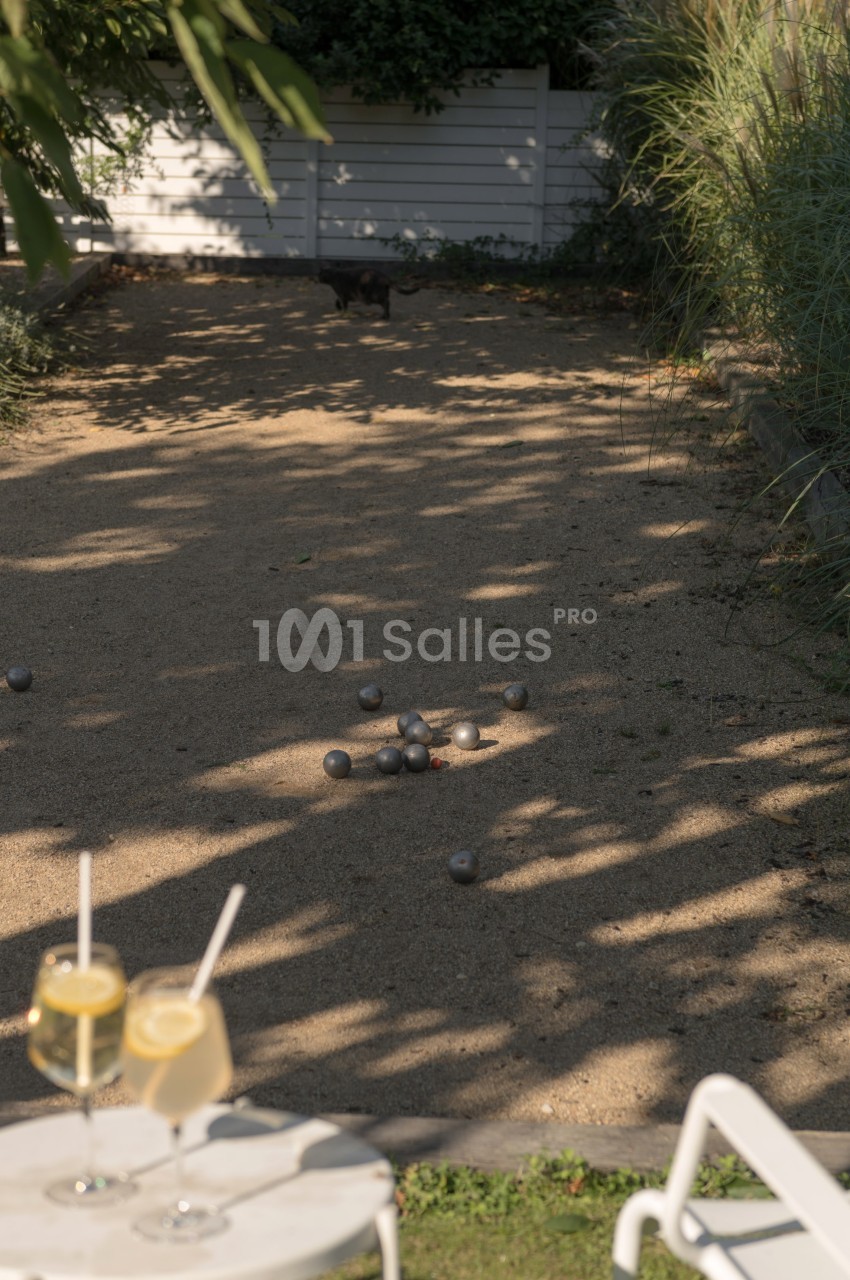 Terrain de pétanque ombragé avec des boules éparpillées au sol, deux verres de boisson posés au premier plan.