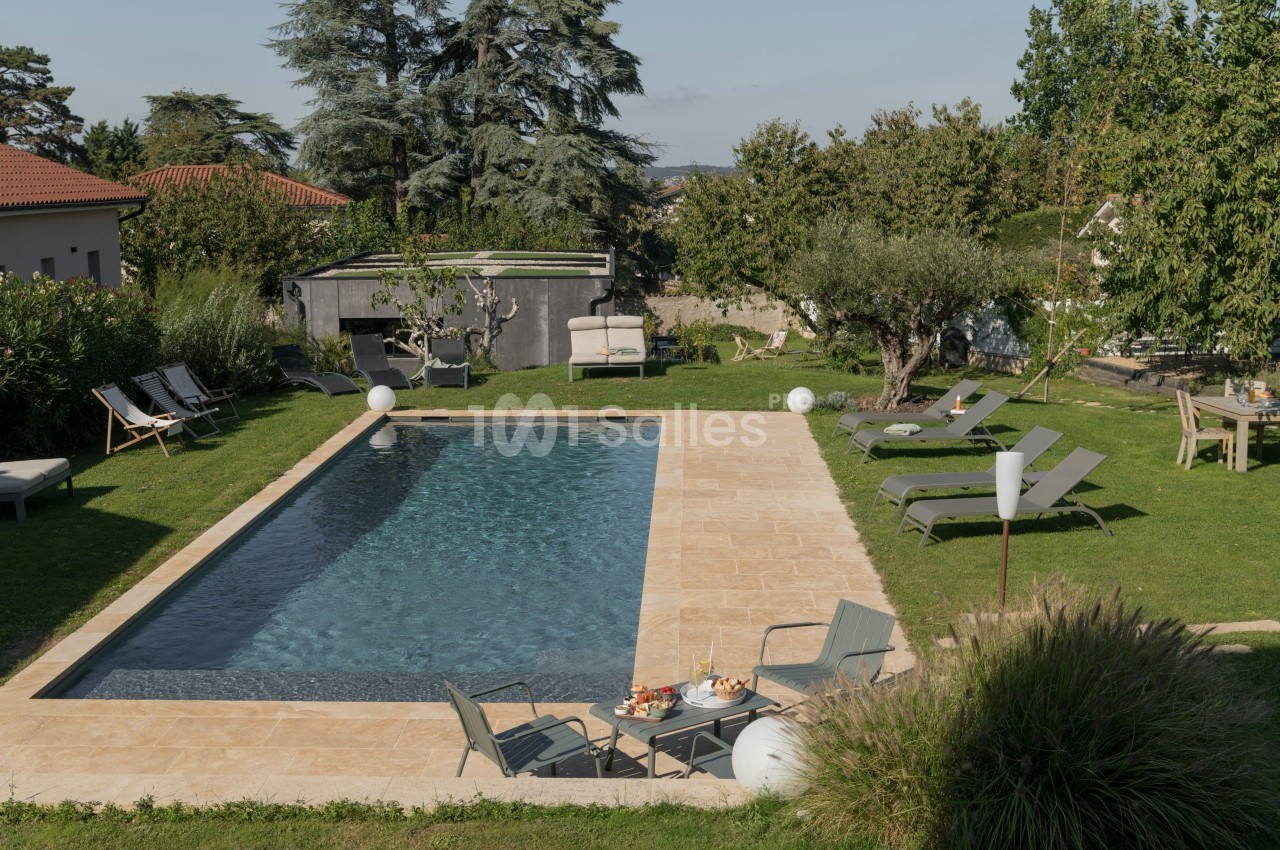 Piscine extérieure entourée de chaises longues, mobilier de jardin et végétation dans un cadre résidentiel.