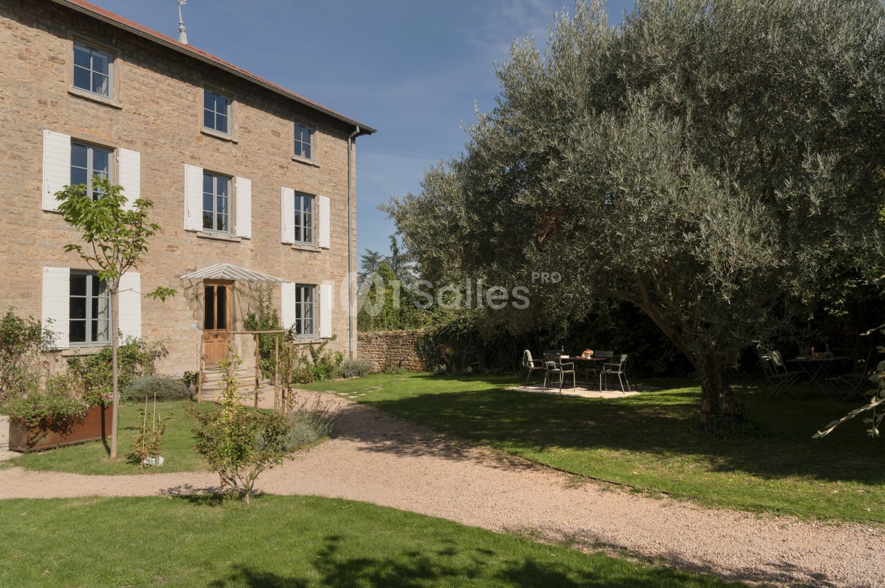 Maison en pierre avec volets blancs entourée d’un jardin arboré, comprenant une terrasse avec table et chaises.