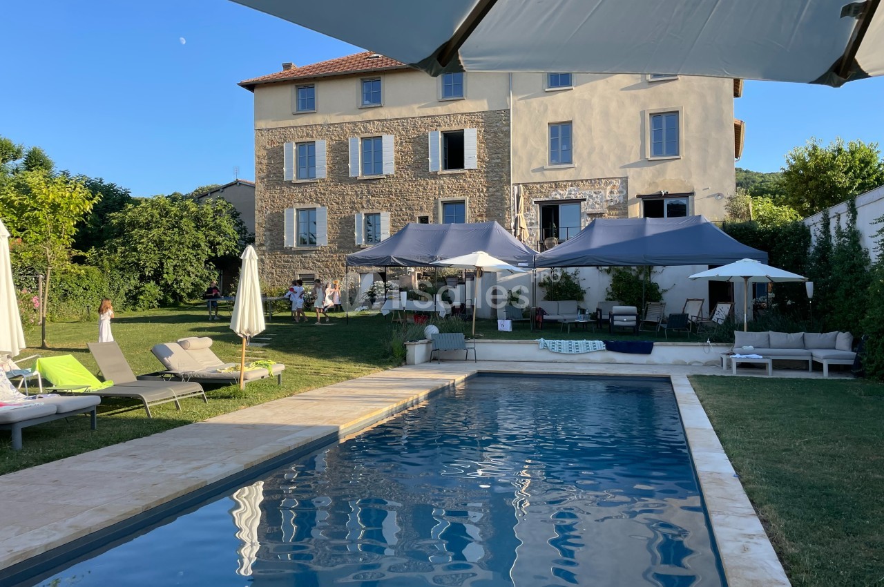 Jardin avec piscine, chaises longues, parasols et grande maison en arrière-plan sous un ciel dégagé.