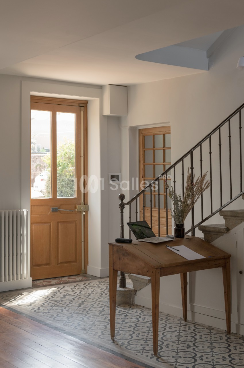 Entrée lumineuse avec porte en bois, table en bois, ordinateur portable et escalier en métal et bois.