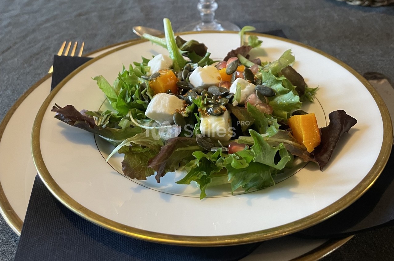 Assiette de salade composée avec feuilles vertes, cubes de fromage, morceaux de potiron et graines, sur une table dressée.
