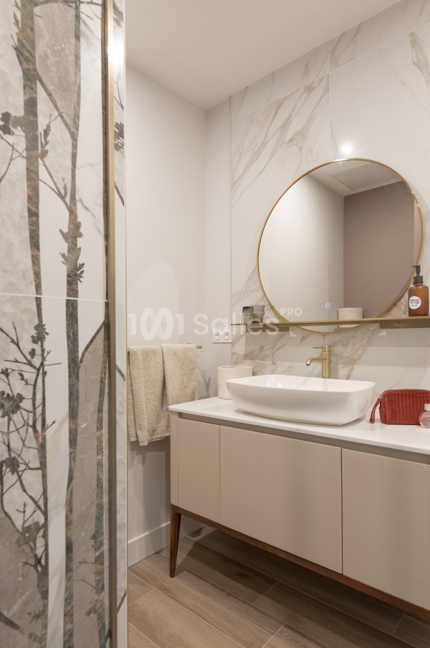 Salle de bain moderne avec lavabo blanc, miroir rond doré, carrelage effet marbre et détails décoratifs naturels.