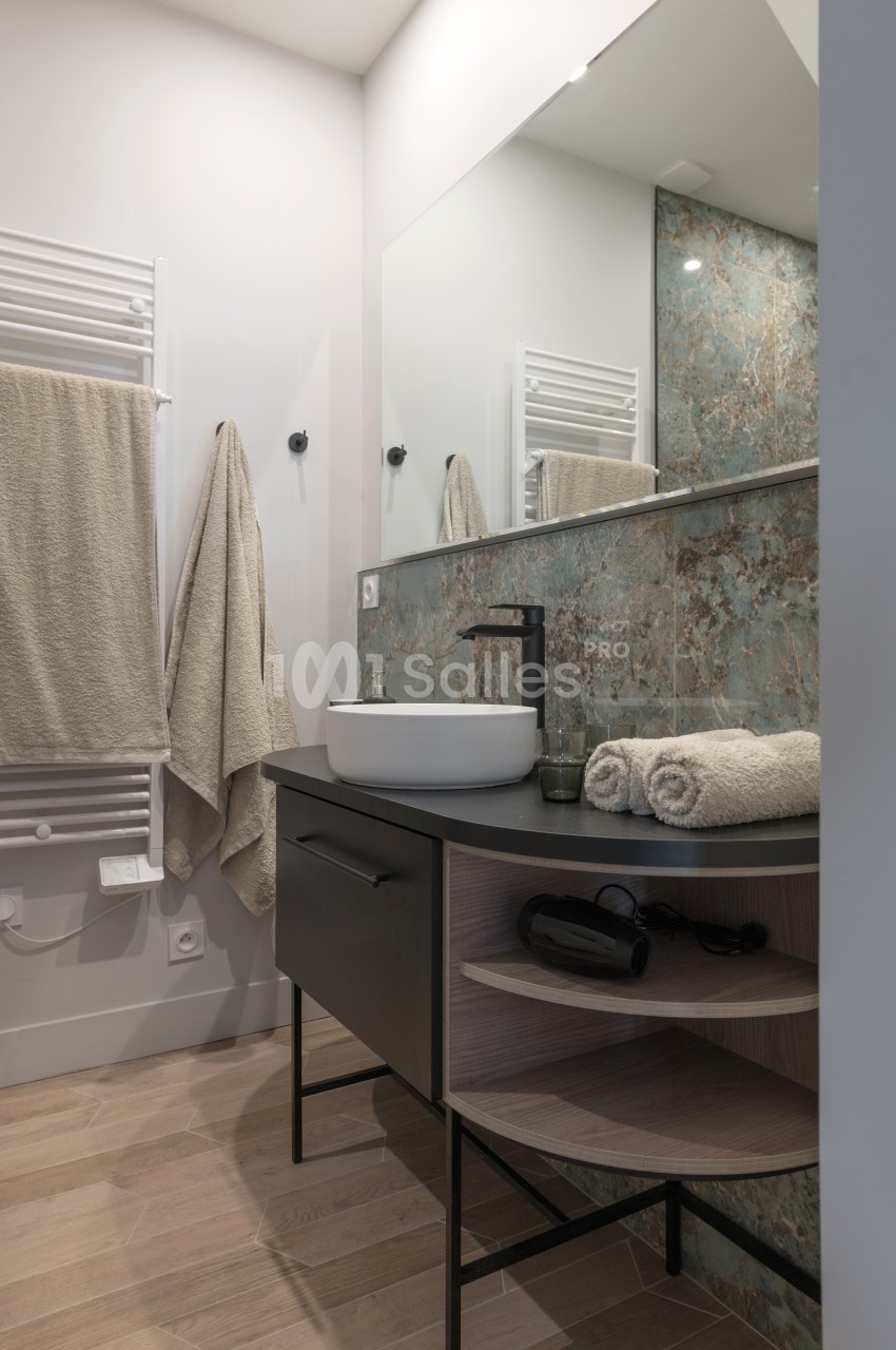 Salle de bain moderne avec lavabo sur meuble noir, miroir, serviettes beiges et mur partiellement carrelé.