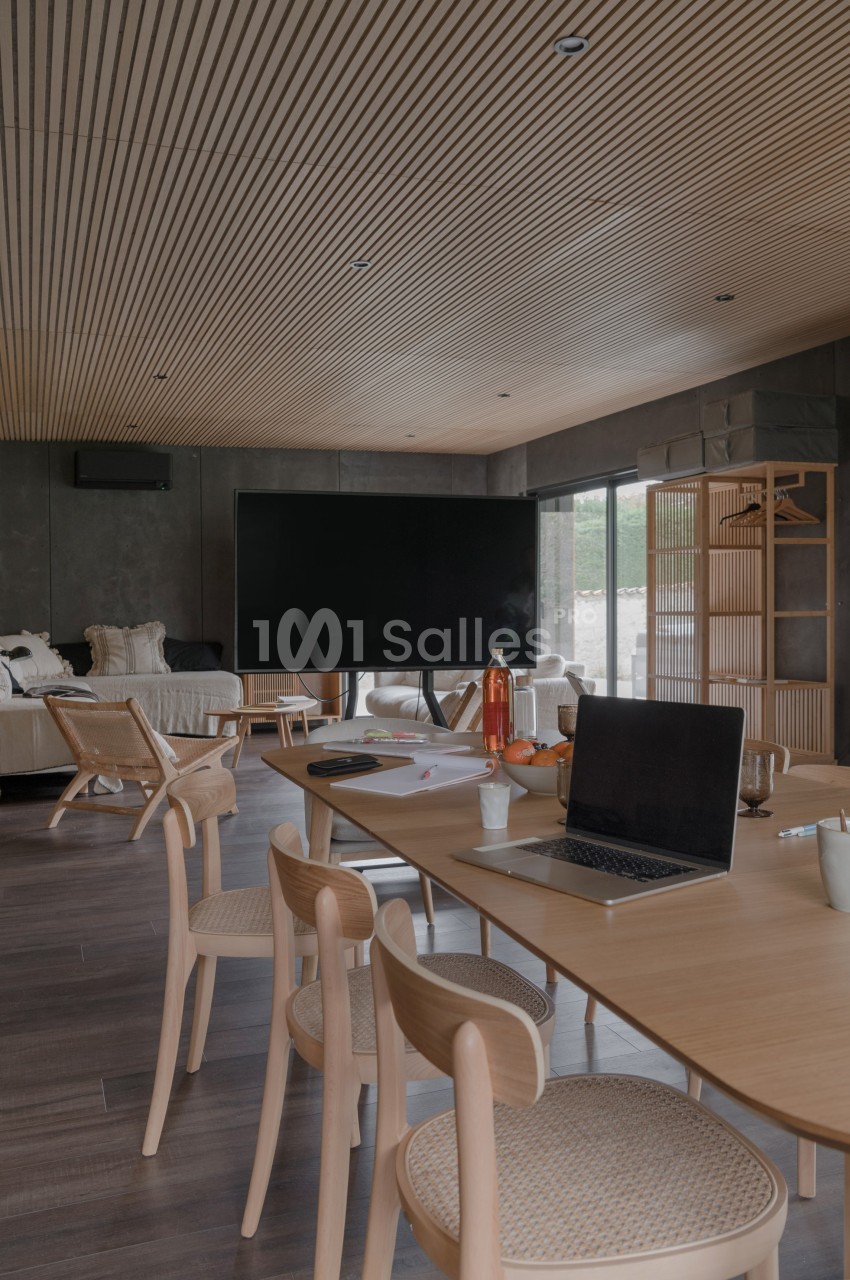 Salle de réunion moderne avec table en bois, chaises, ordinateur portable et écran noir, éclairée par une lumière naturelle.