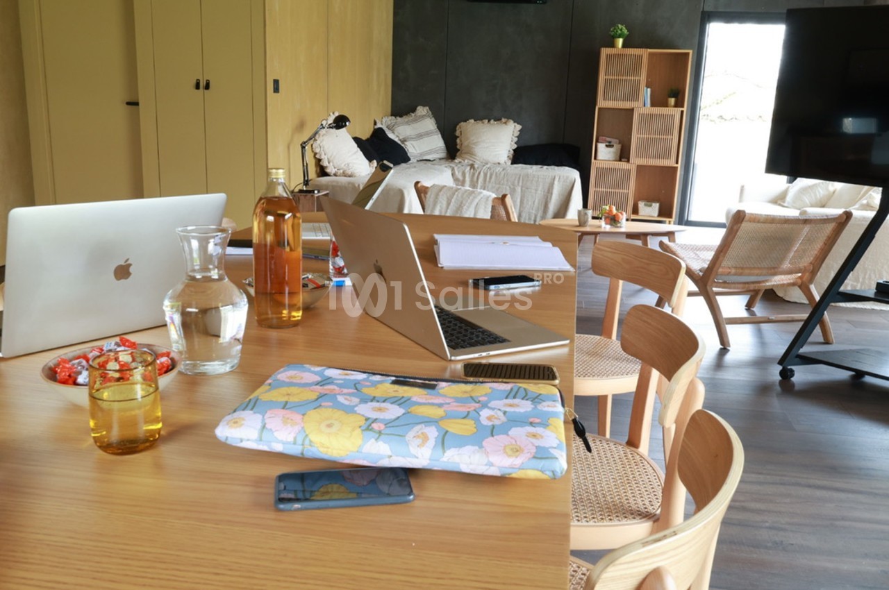 Table en bois avec ordinateurs portables, bouteille, verre d'eau et objets divers dans un salon lumineux et moderne.