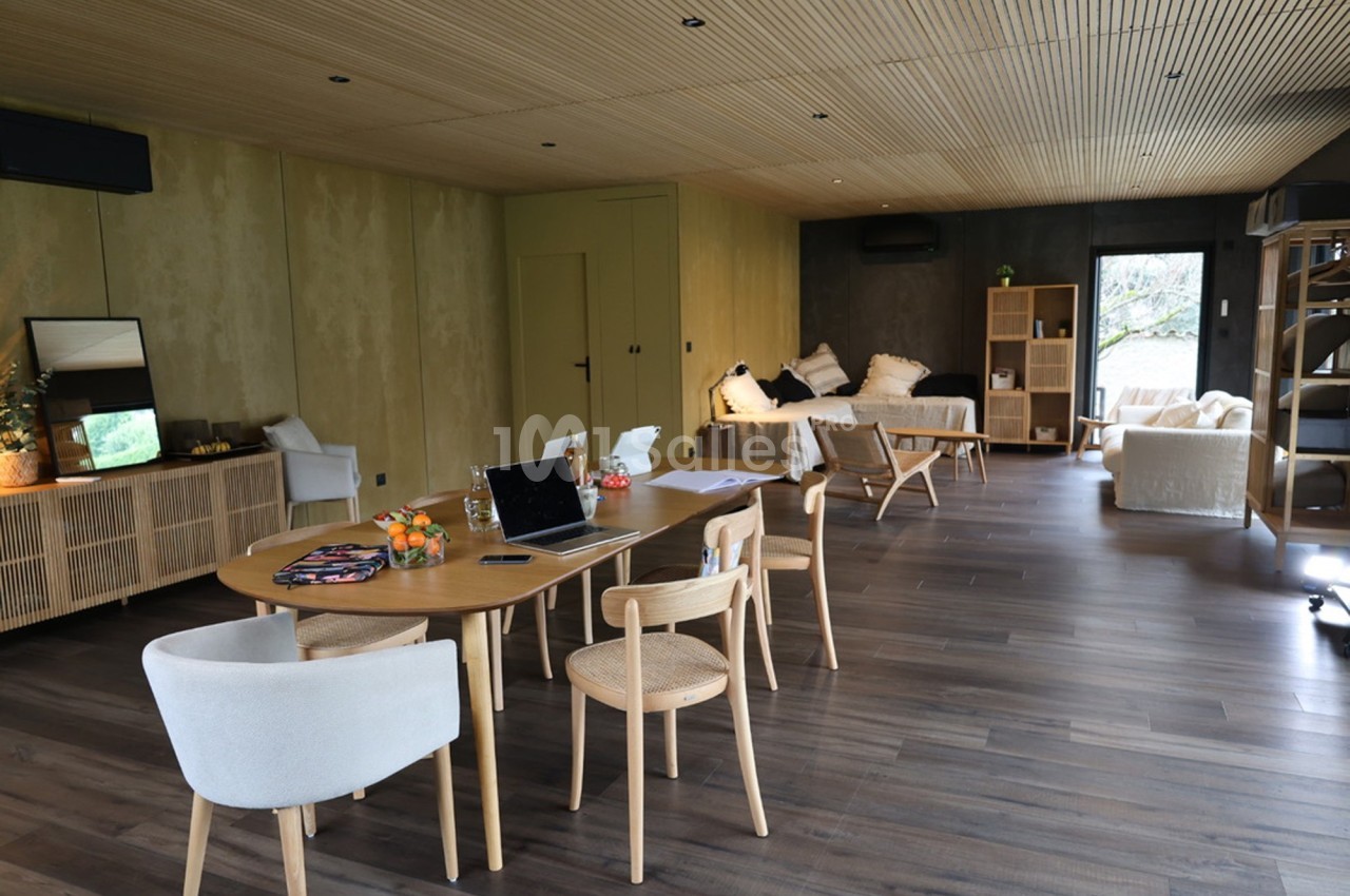Salle lumineuse avec parquet foncé, mobilier en bois clair, table de travail, canapé et grandes baies vitrées.