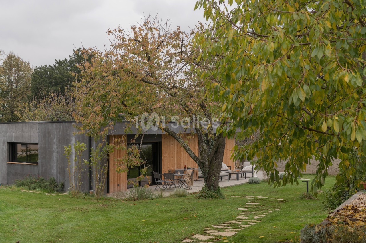 Maison contemporaine en bois et béton entourée d'un jardin verdoyant avec un arbre au premier plan.