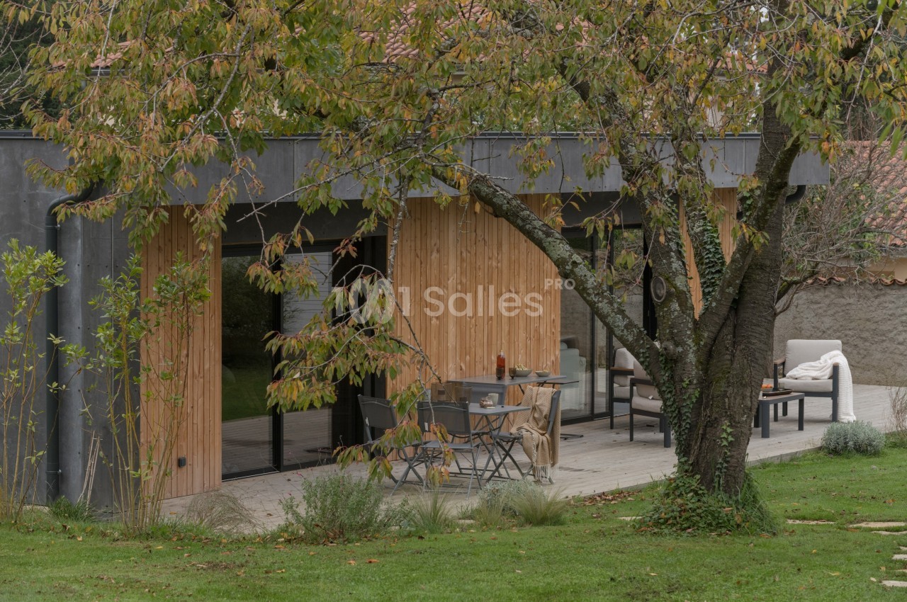 Terrasse en bois aménagée avec table et chaises, entourée de verdure et d'un arbre aux feuilles automnales.