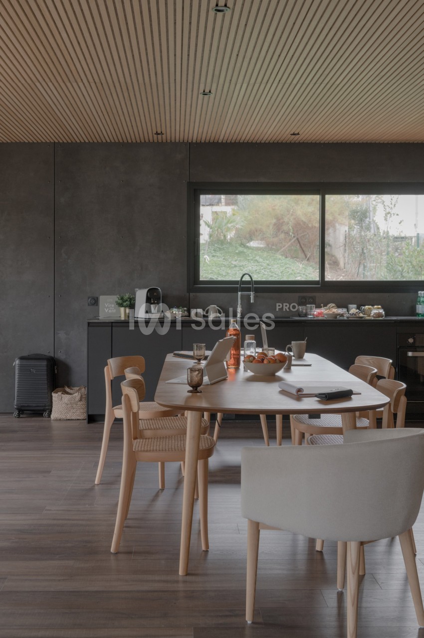 Salle à manger moderne avec table en bois, chaises assorties, cuisine ouverte et grande fenêtre donnant sur un jardin.
