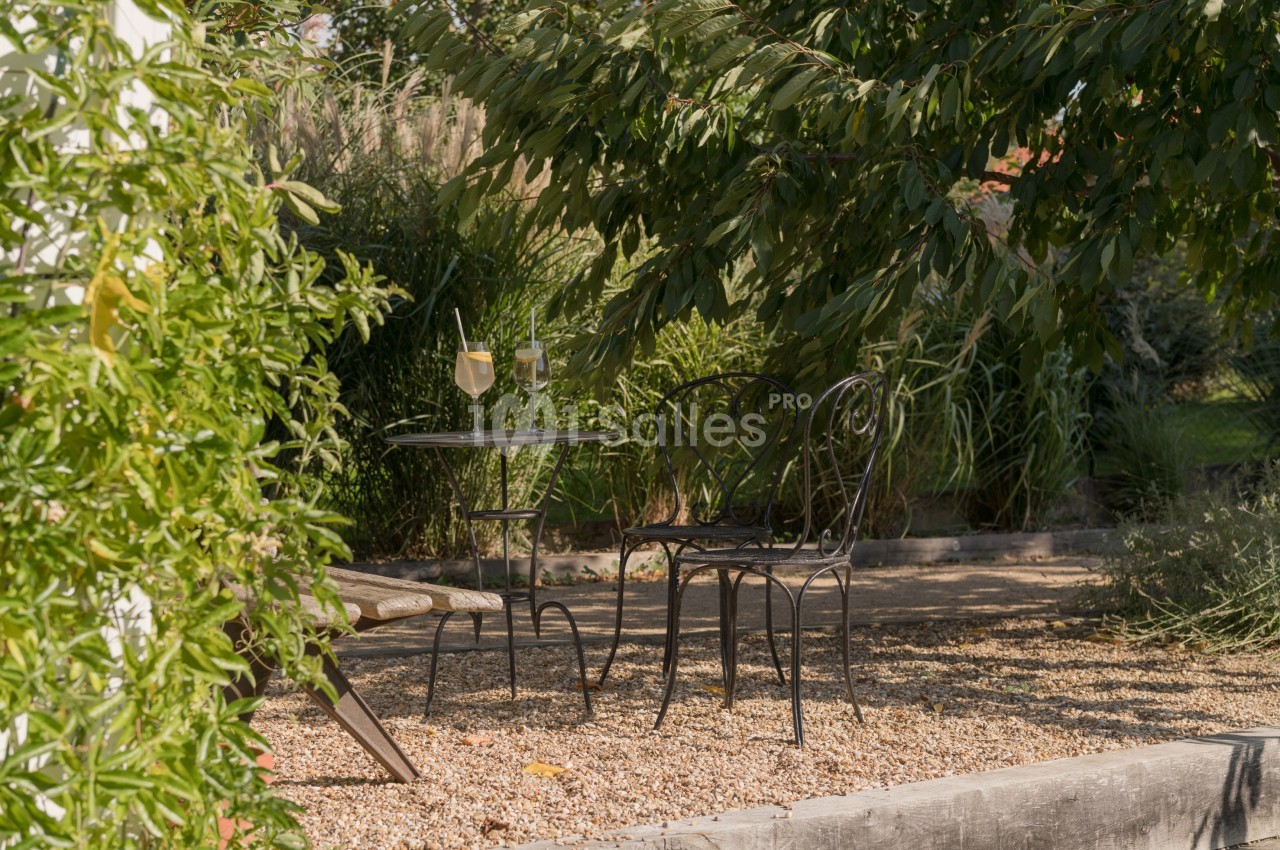 Table et chaises en fer forgé sous un arbre, avec deux verres posés, dans un jardin ombragé sur sol gravillonné.