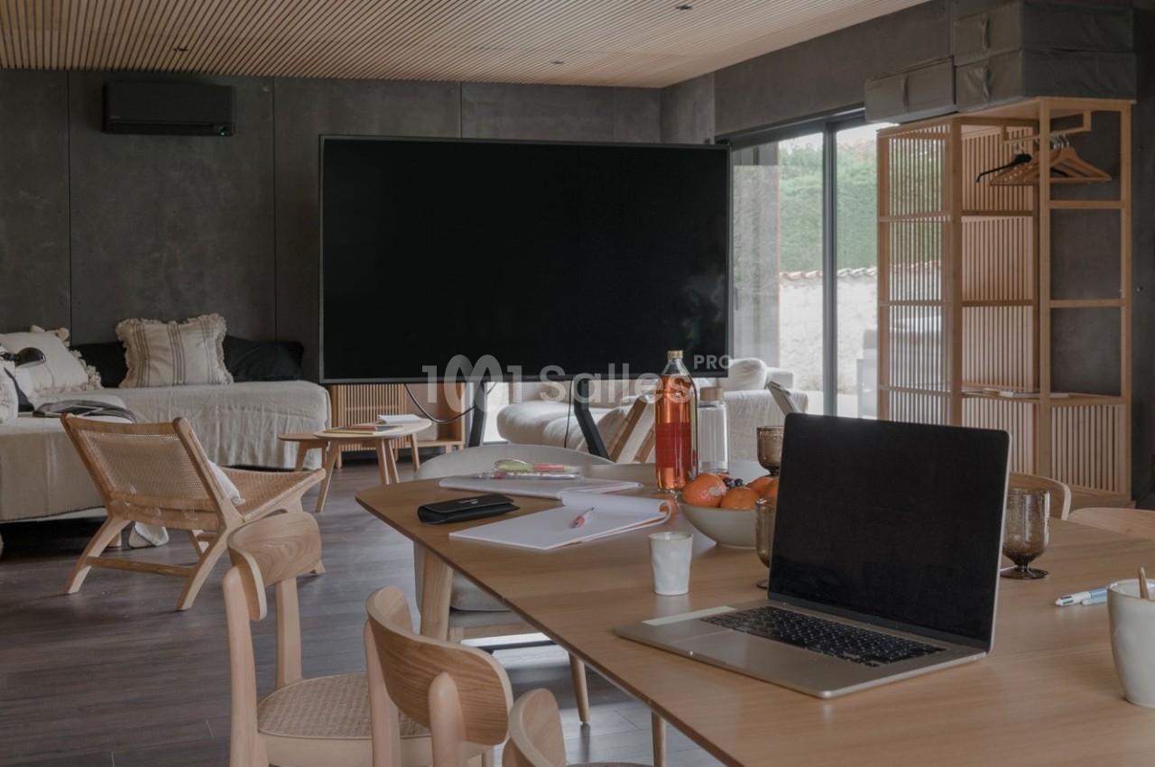 Espace de travail dans un salon moderne avec table en bois, ordinateur portable, écran, et lumière naturelle.