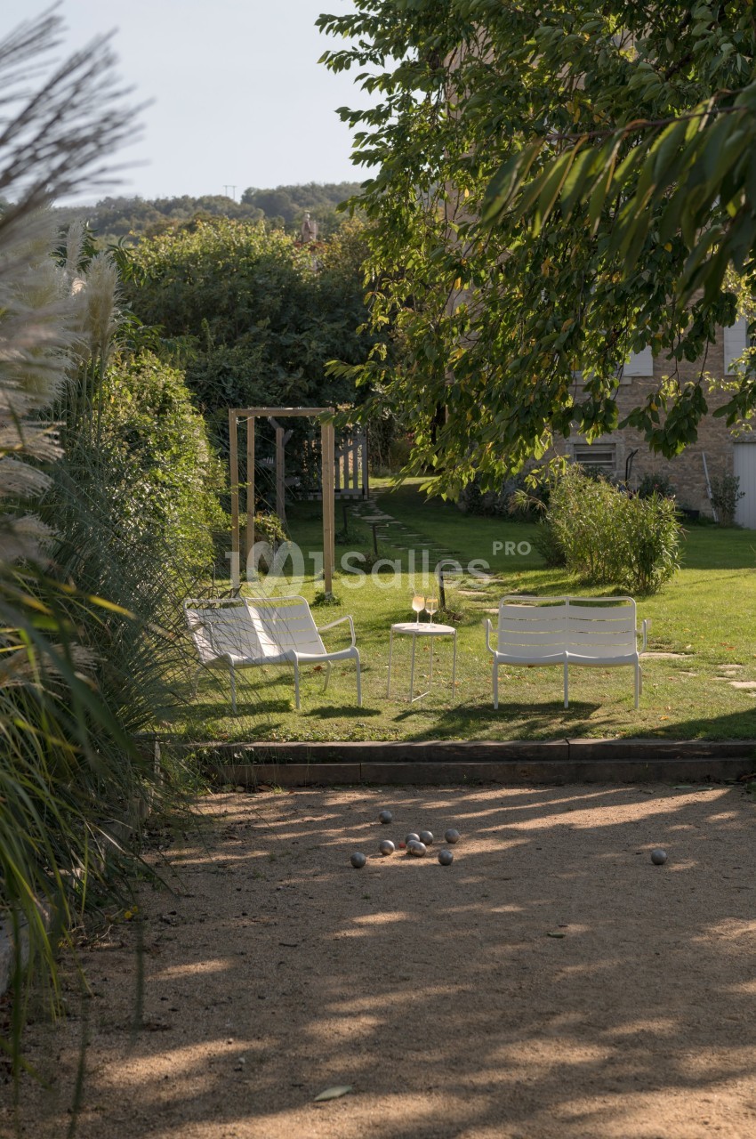 Deux chaises blanches et des verres de vin sur une table dans un jardin, avec un terrain de pétanque au premier plan.