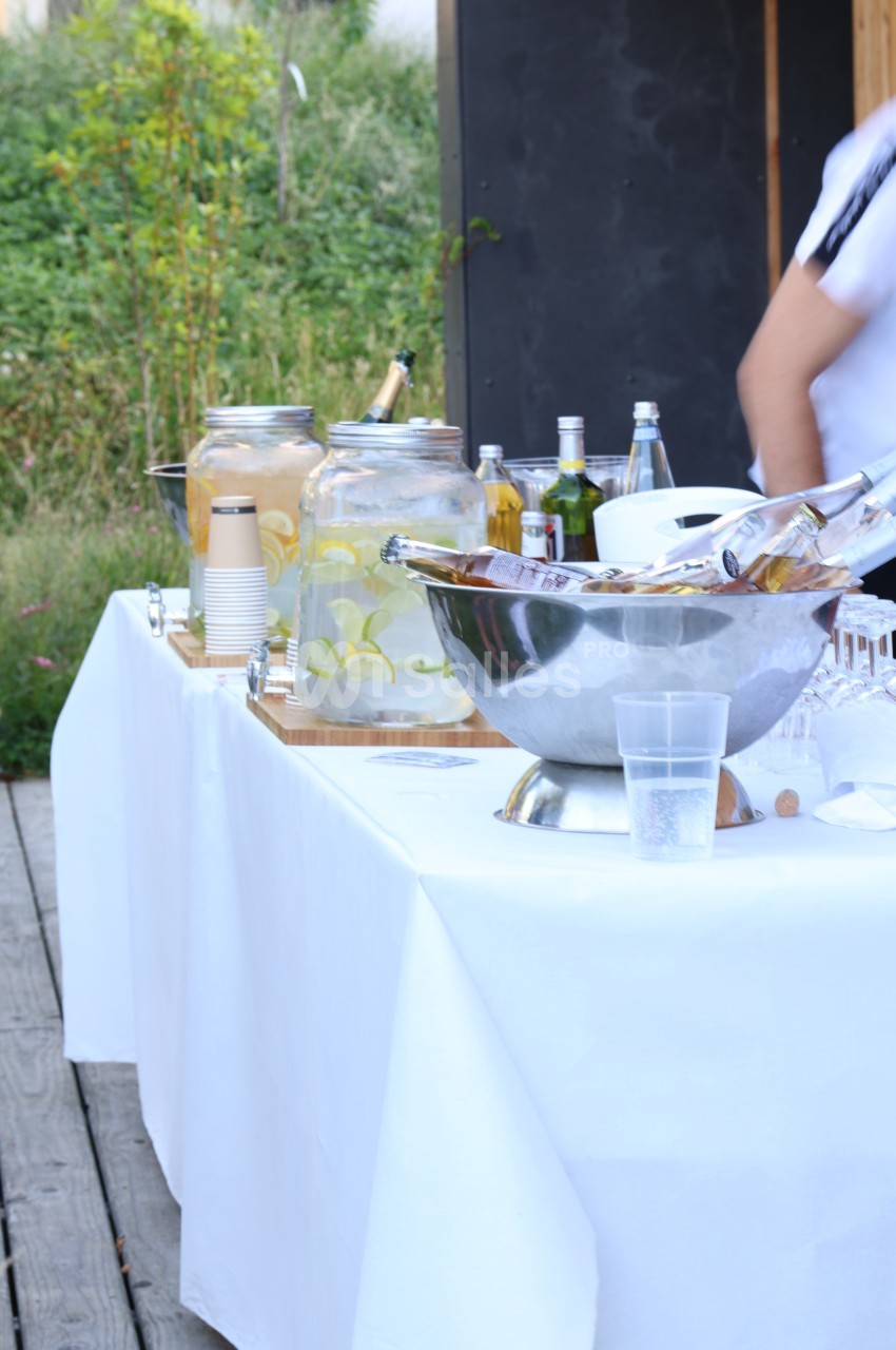 Table dressée en extérieur avec boissons fraîches, carafes d'eau infusée, bouteilles et verres sur une nappe blanche.