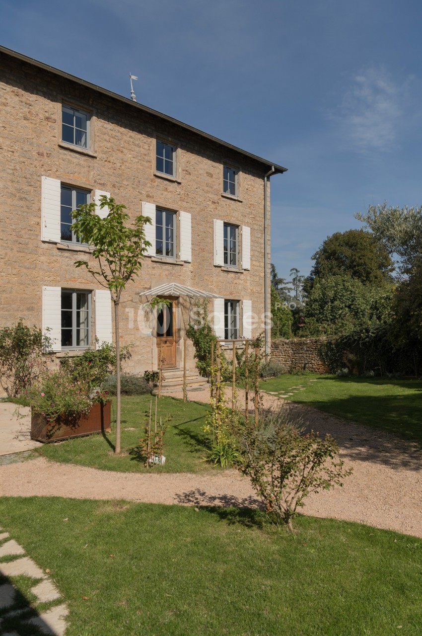 Façade en pierre d'une maison à trois étages avec volets blancs, jardin arboré et allée gravillonnée au premier plan.