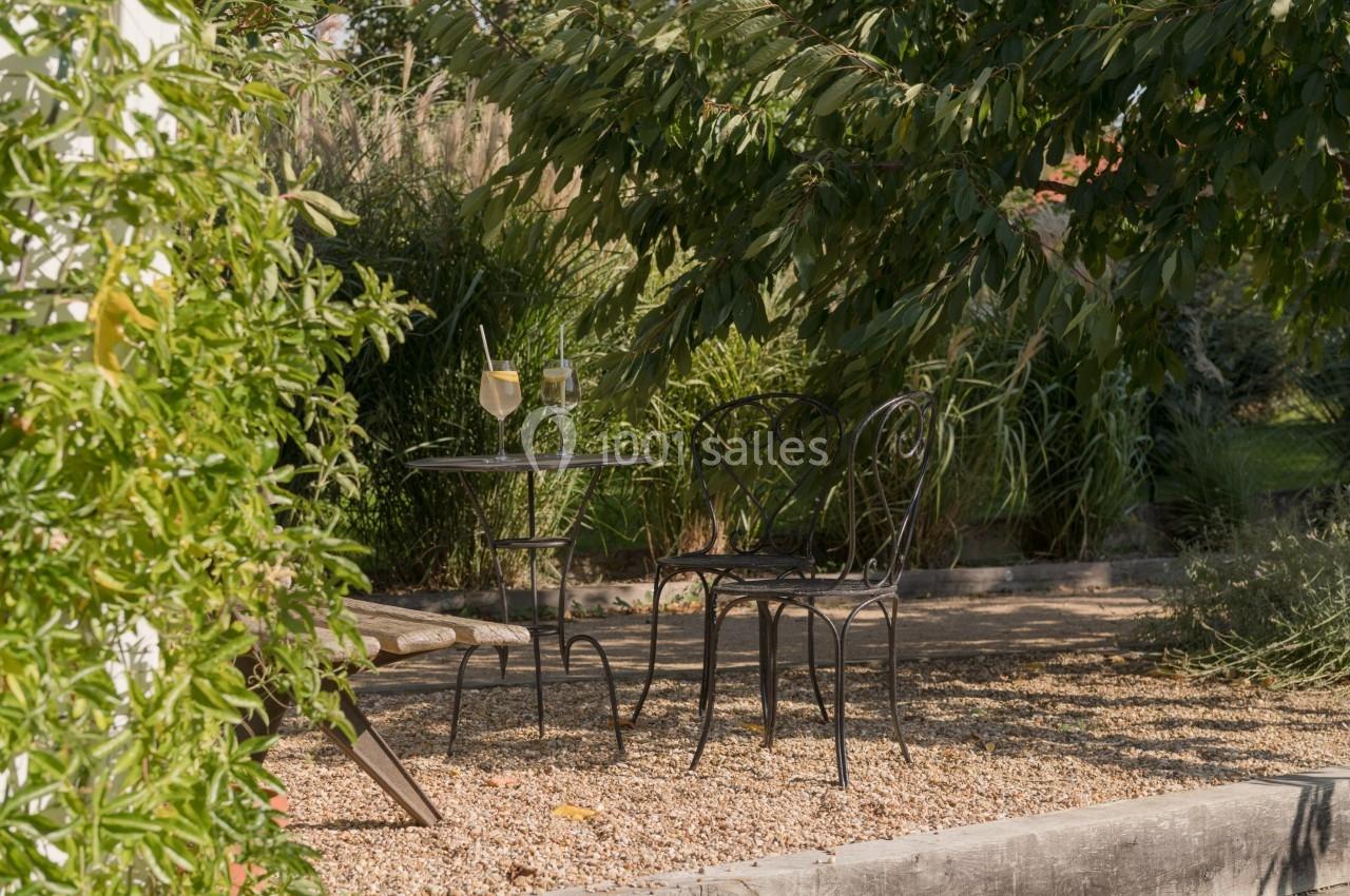 Table et chaises en fer forgé sous des arbres, avec des verres posés dessus, dans un jardin ombragé sur sol gravillonné.