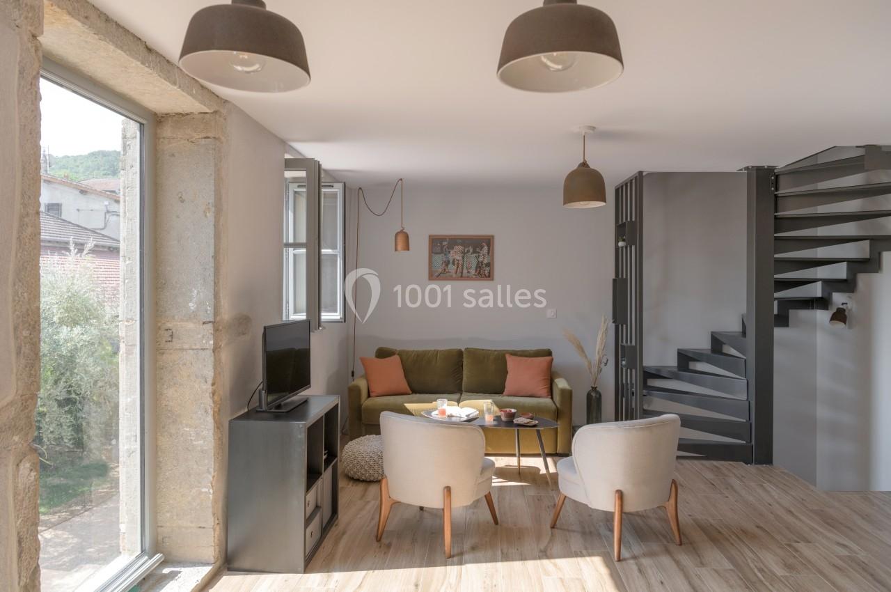 Salon lumineux avec canapé vert, deux fauteuils, table basse, escalier métallique et grande fenêtre donnant sur l'extérieur.