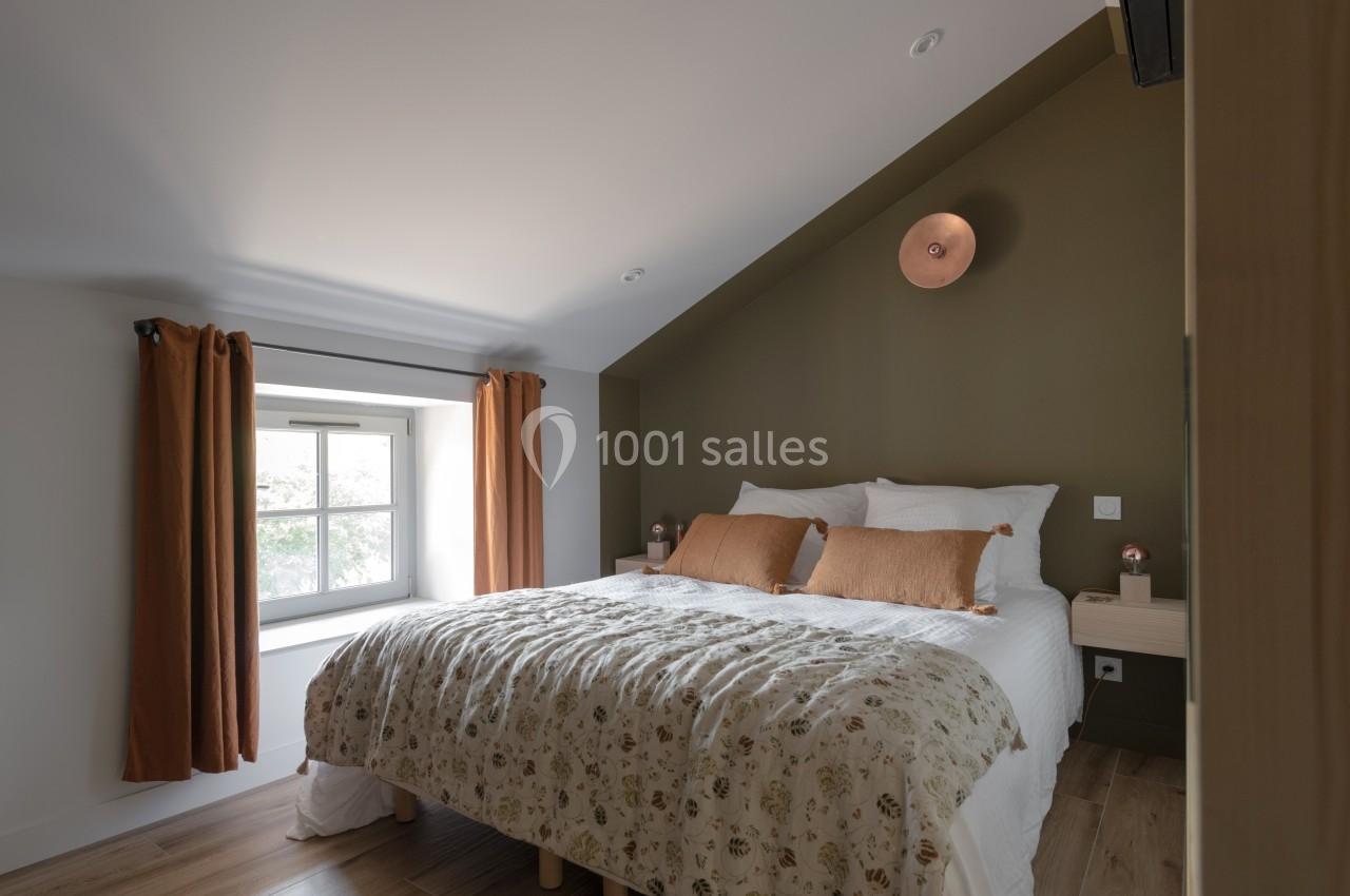 Chambre lumineuse avec un lit double, des coussins orange, un mur vert olive et une fenêtre avec rideaux assortis.