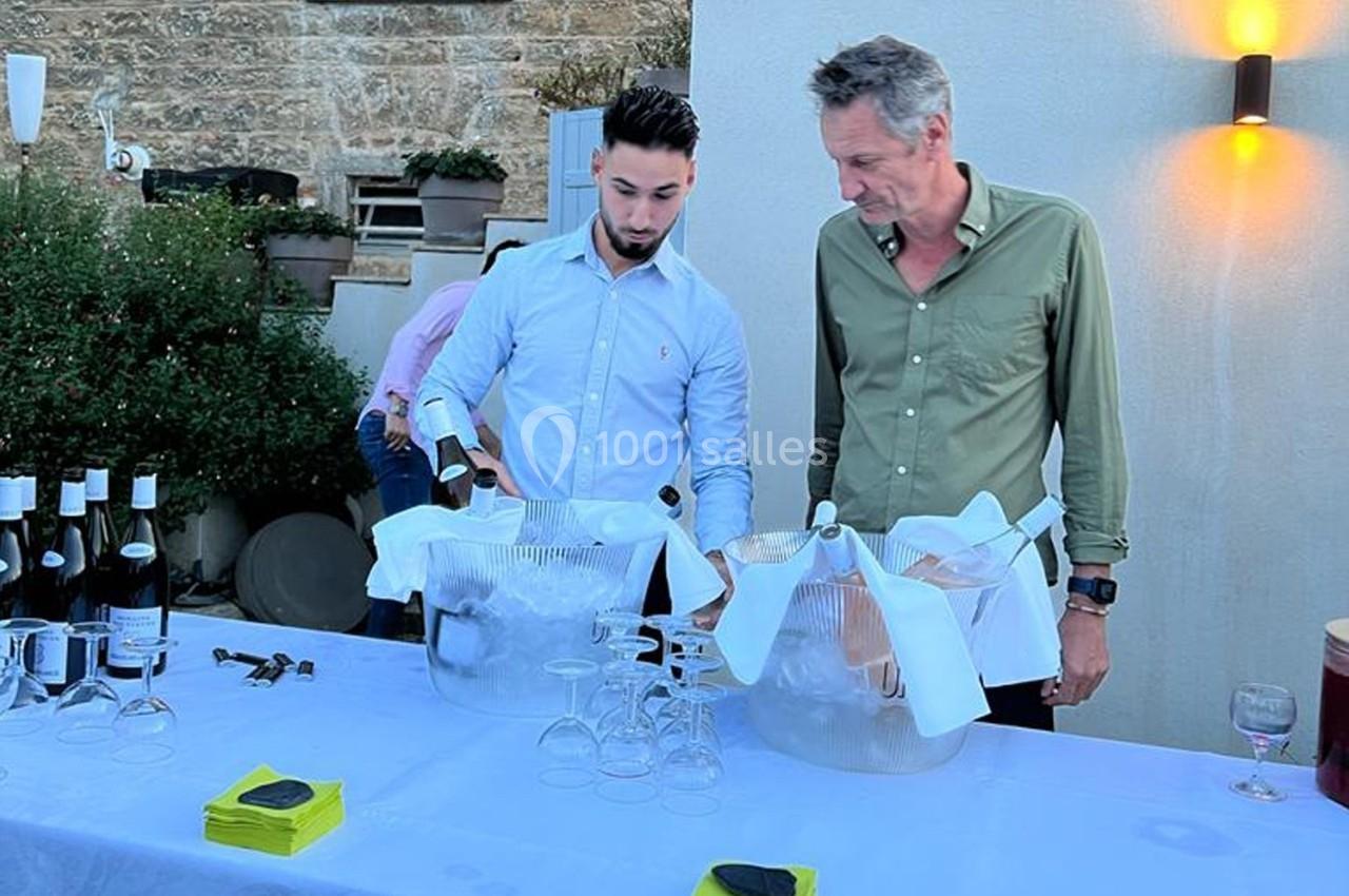 Deux hommes préparent des bouteilles de vin dans des seaux à glace sur une table dressée en extérieur.