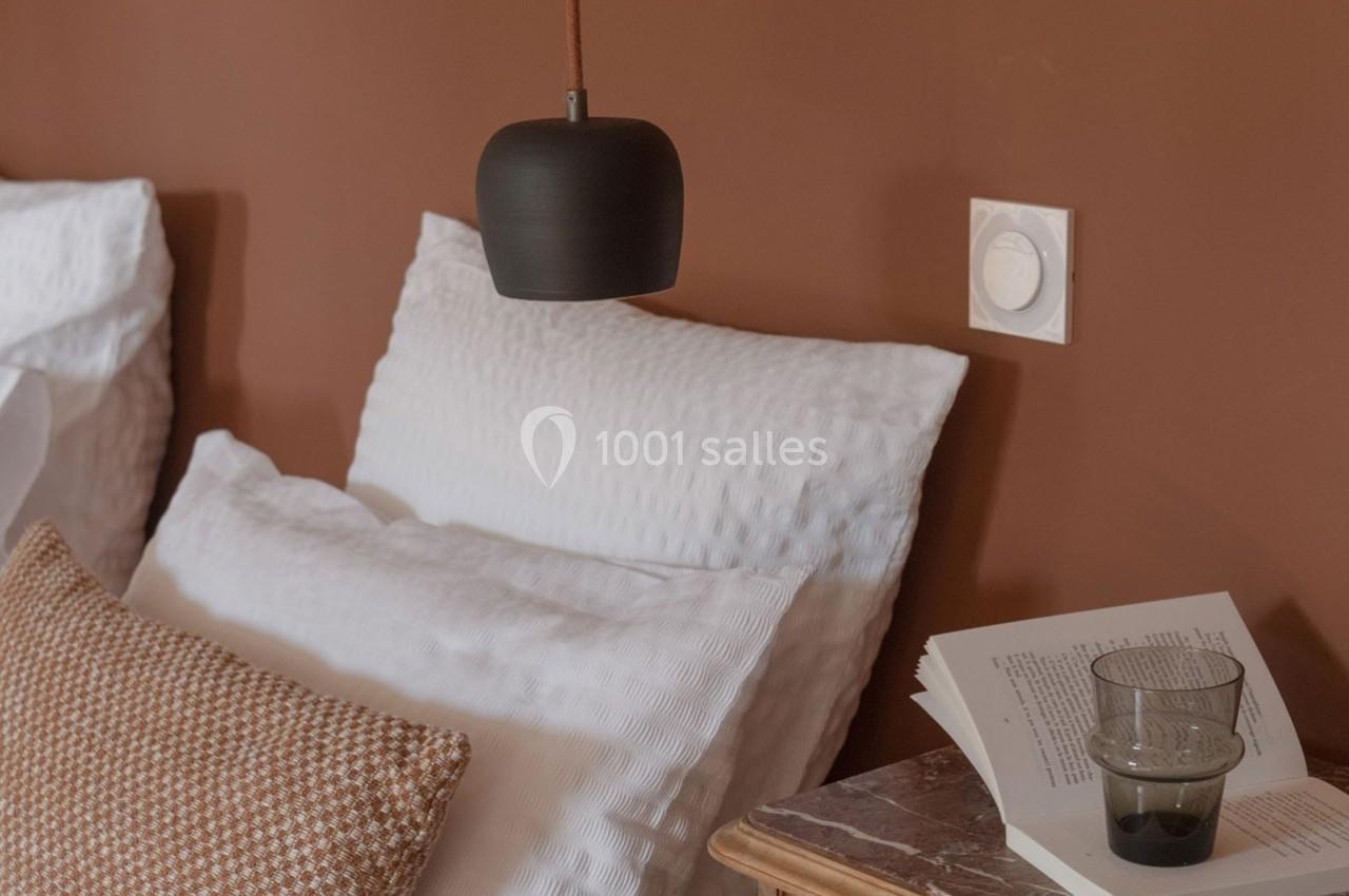 Lampe suspendue noire au-dessus d'un lit avec oreillers blancs, table de chevet en bois, livre ouvert et verre d'eau.