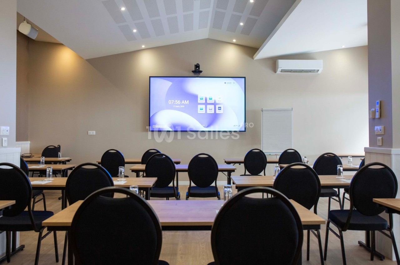 Salle de réunion équipée de tables, chaises, écran de projection allumé, climatisation et éclairage au plafond.