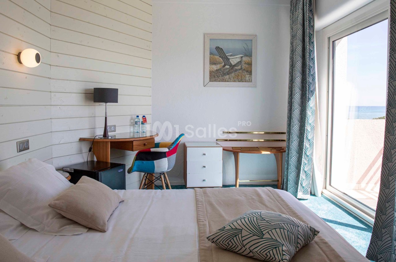 Chambre lumineuse avec lit double, bureau en bois, fauteuil coloré et grande baie vitrée donnant sur la mer.