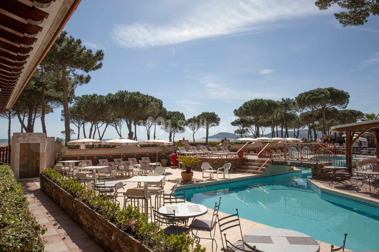 Piscine extérieure entourée de chaises longues et de tables, avec des pins et un ciel dégagé en arrière-plan.
