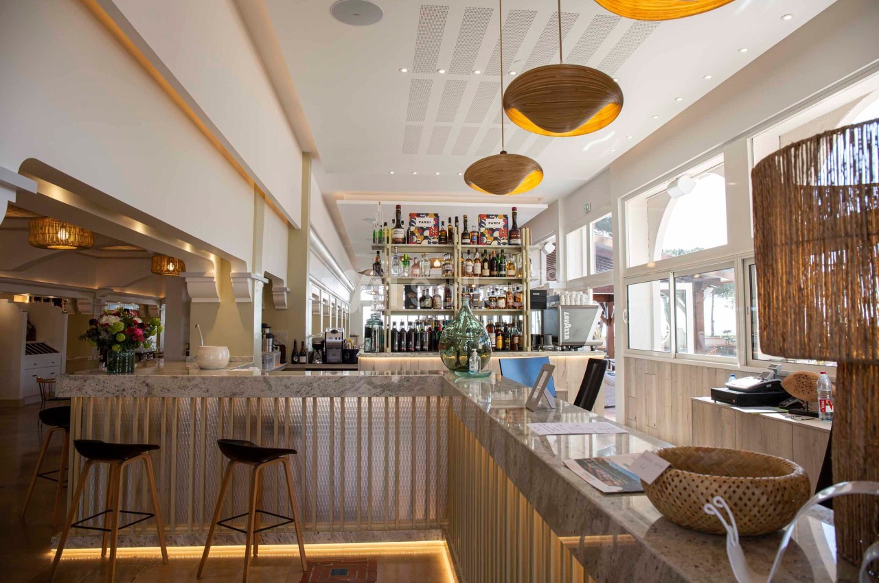 Intérieur d'un bar lumineux avec comptoir en marbre, étagères de bouteilles, tabourets et décorations en bois.