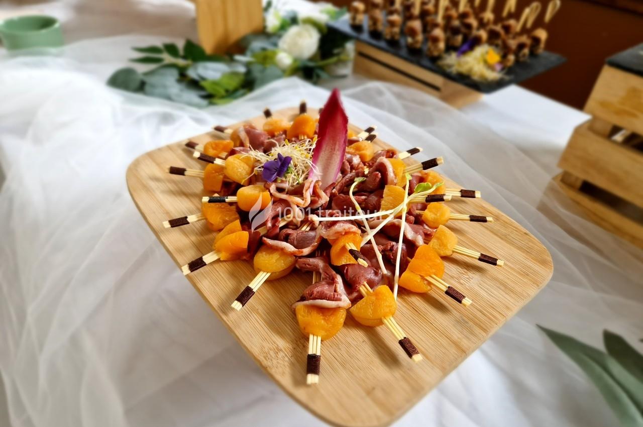 Plateau de brochettes apéritives avec morceaux de viande, quartiers d'abricots et décoration florale sur une table.