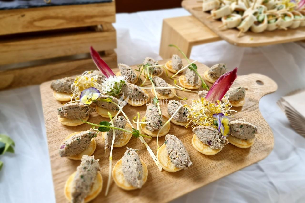 Plateau de mini-blinis garnis de rillettes et décorés de pousses et fleurs comestibles, présenté sur une planche en bois.