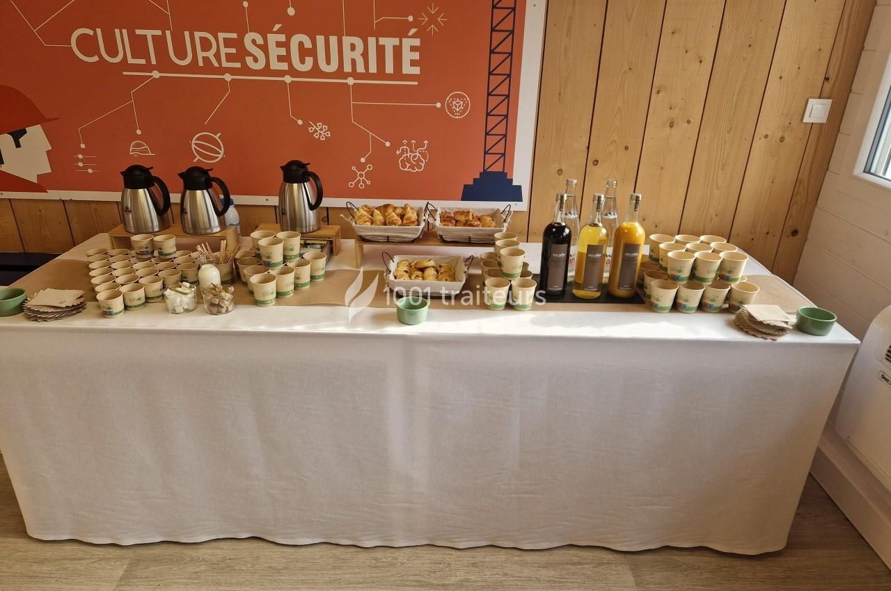 Table dressée avec boissons chaudes, jus, viennoiseries et gobelets, dans une salle avec un panneau ’Culture Sécurité’.