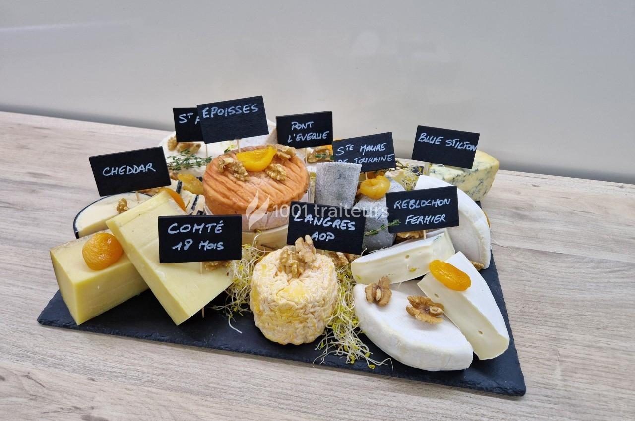 Plateau de fromages variés sur une ardoise, avec étiquettes indiquant les noms, décoré de fruits secs et herbes.