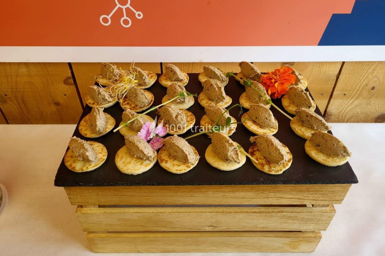Plateau de mini-blinis garnis de mousse de foie et décorés de fleurs comestibles, présenté sur une ardoise.