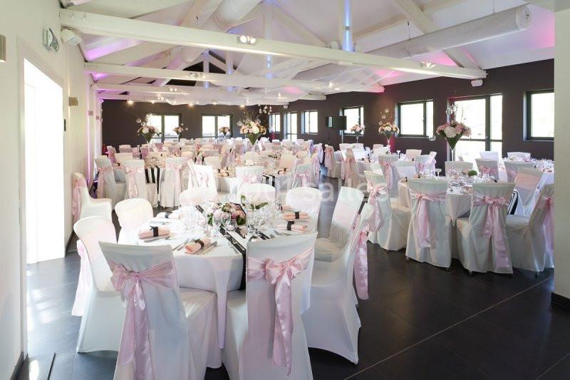 Salle de réception décorée pour un mariage avec des tables rondes, nappes blanches et chaises ornées de rubans roses.