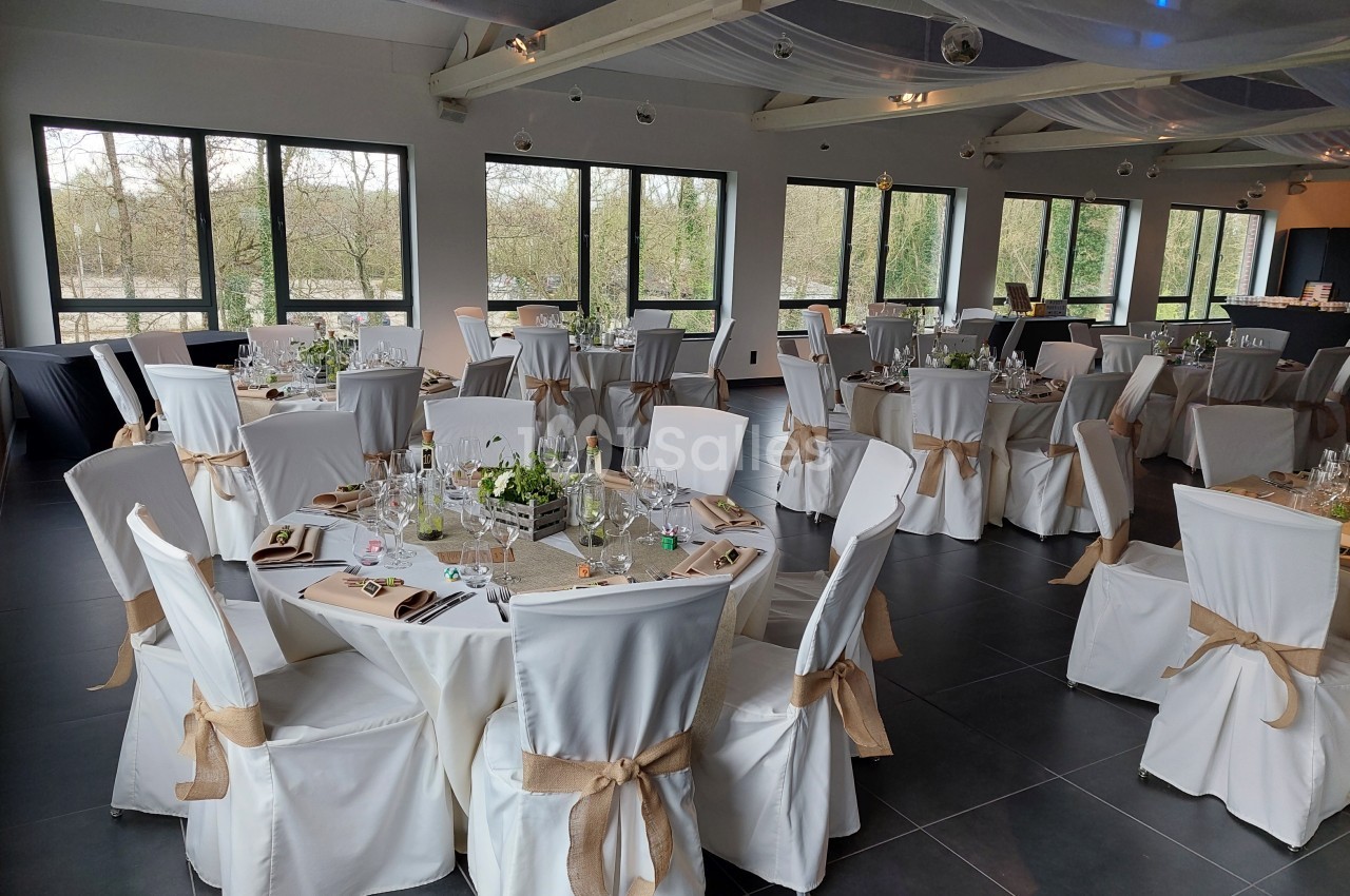 Salle de réception décorée avec des tables rondes dressées, nappes blanches et chaises ornées de nœuds en toile de jute.