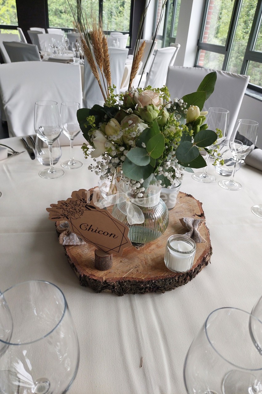 Centre de table avec fleurs blanches et feuillages dans un vase, posé sur une tranche de bois avec une bougie.