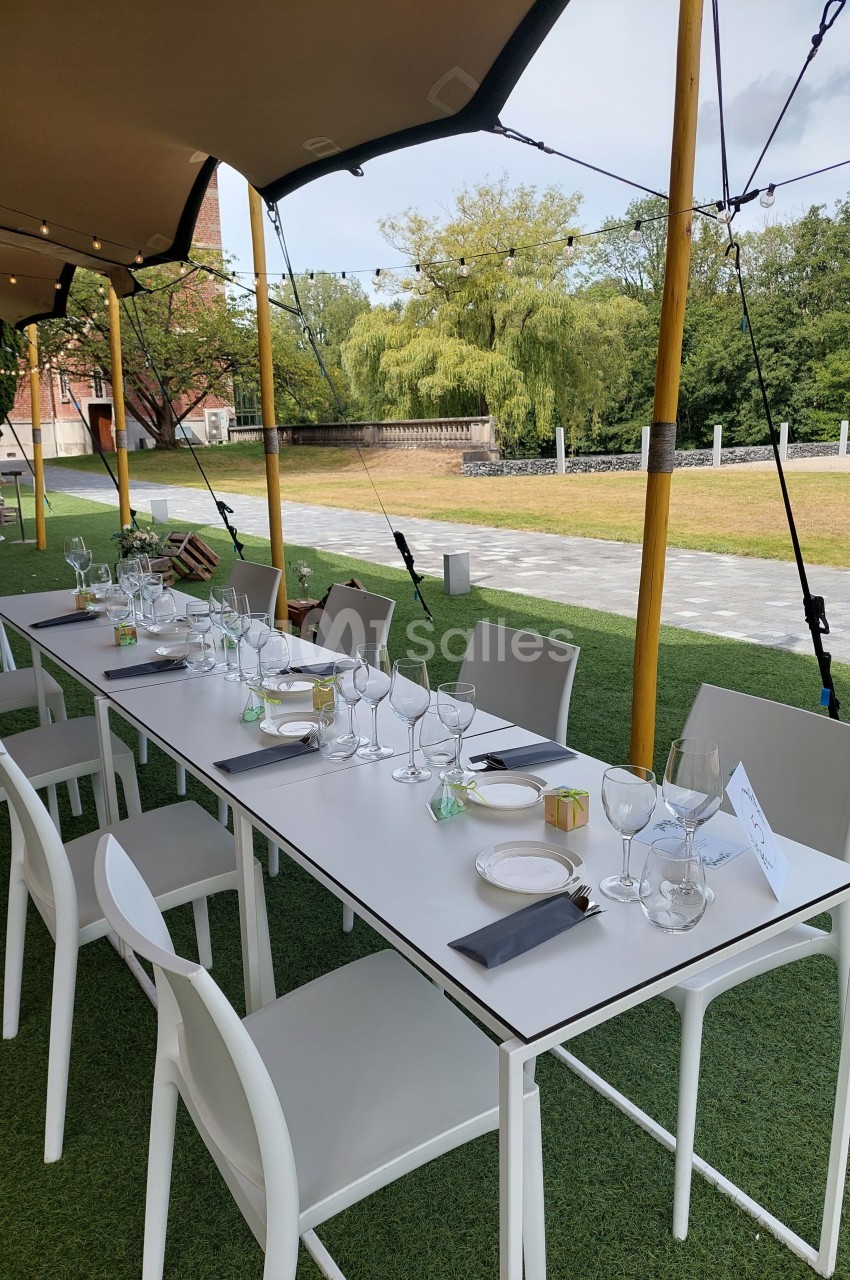 Table dressée en extérieur sous une tente, avec chaises blanches, verres, couverts et serviettes sur une pelouse.