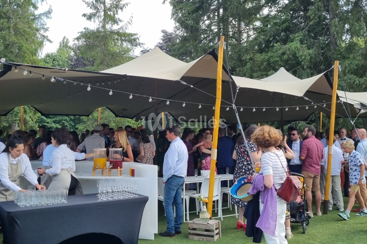 Groupe de personnes réunies sous une tente dans un jardin, avec des tables de service et des décorations lumineuses.