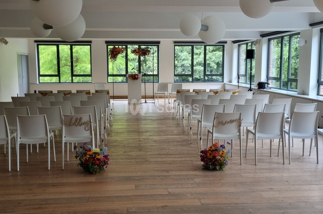 Salle lumineuse aménagée pour une cérémonie, avec des chaises blanches alignées et des bouquets de fleurs colorées.