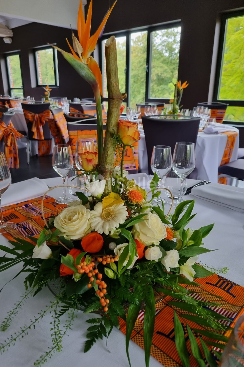 Centre de table floral avec roses, gerberas, feuillage et oiseaux de paradis, dans une salle de réception lumineuse.