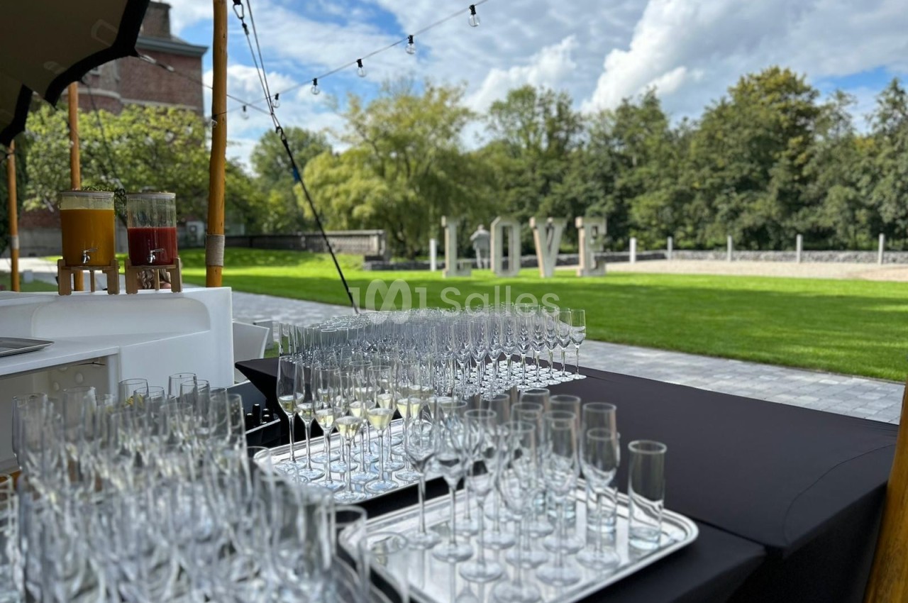 Verres à pied alignés sur des plateaux sur une table noire en extérieur, avec un jardin et des arbres en arrière-plan.