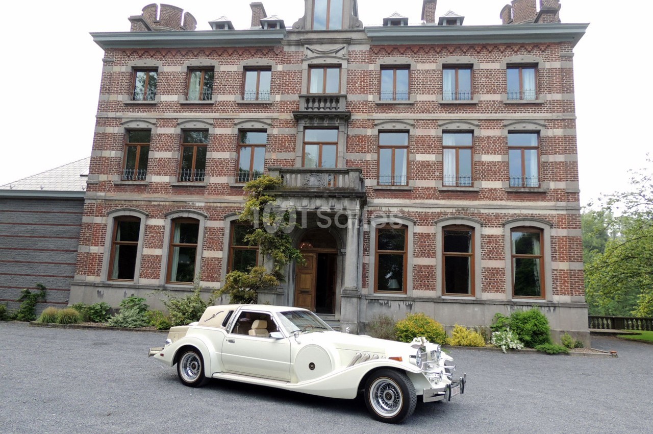 Voiture ancienne blanche garée devant une grande maison en briques rouges avec des fenêtres symétriques.