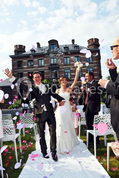 Un couple souriant marche sur une allée extérieure décorée, entouré d'invités lançant des pétales de fleurs.