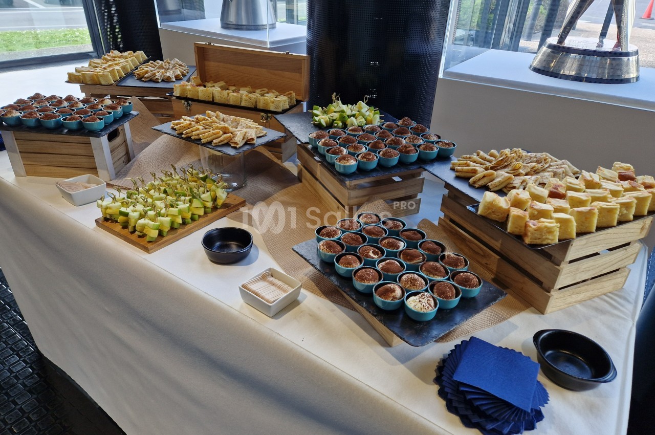 Buffet composé de verrines, amuse-bouches et petits fours disposés sur des présentoirs en bois dans une salle lumineuse.